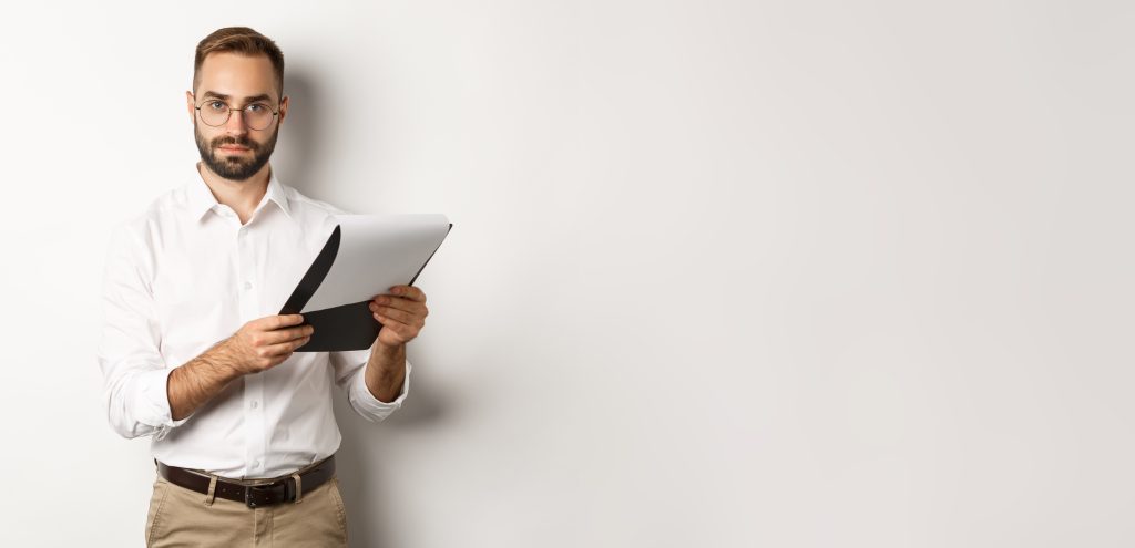 serious employer looking at camera while reading documents on clipboard, having job interview, standing over white background