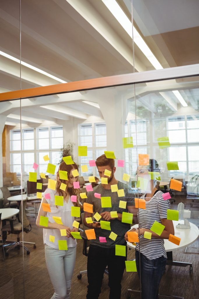 business executives standing near glass with sticky notes and discussing over digital tablet